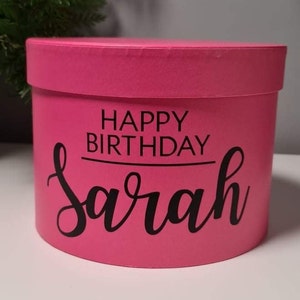 Pu&ograve; includere: Una scatola regalo rotonda rosa con il testo "Happy Birthday Sarah" in lettere nere.
