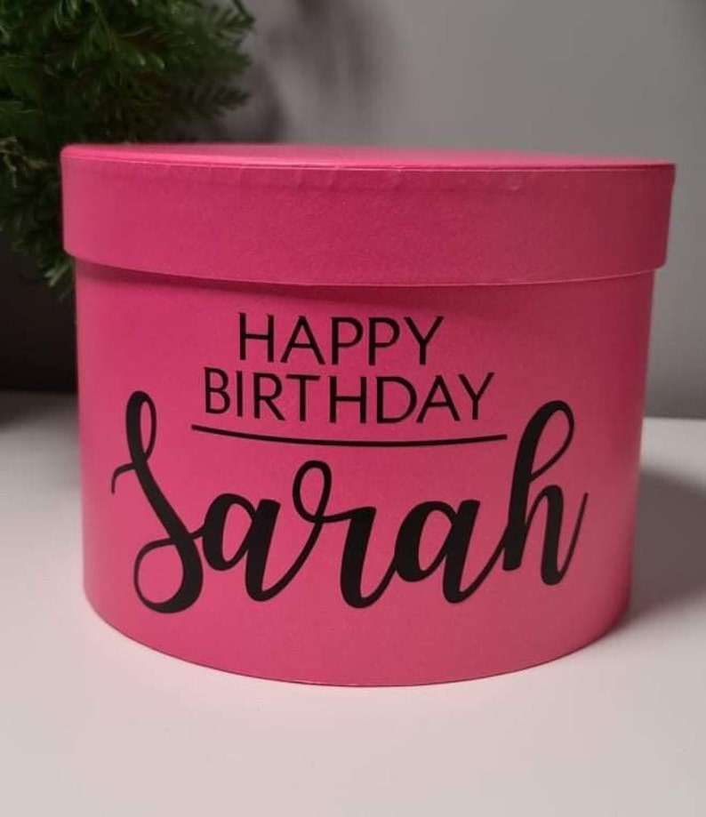 Pu&ograve; includere: Una scatola regalo rotonda rosa con il testo "Happy Birthday Sarah" in lettere nere.