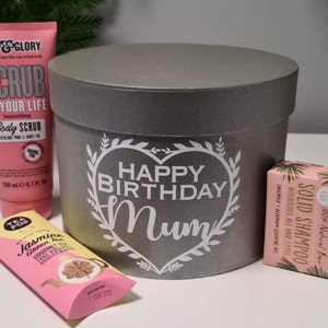 Pu&ograve; includere: Una scatola regalo rotonda grigia con un design a forma di cuore bianco che dice "Happy Birthday Mum". La scatola &egrave; circondata da una variet&agrave; di regali, tra cui uno scrub corpo rosa, una scatola di bustine di t&egrave; rosa e una saponetta rosa.