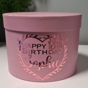 Pu&ograve; includere: Una scatola regalo rotonda rosa con un design in foglia d'oro che dice "Happy 18th Birthday Sophie" a forma di cuore.