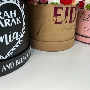Pode incluir: Tr&ecirc;s caixas de presente redondas com arranjos florais. A caixa preta tem um cora&ccedil;&atilde;o com o texto "Umrah Ubarak Sonia" e as palavras "Forts and Bless You". A caixa marrom tem o texto "Eid Mubarak" em dourado e marrom. A caixa rosa tem o texto "Eid Mubarak Aysha" em preto.