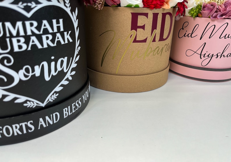 Pode incluir: Tr&ecirc;s caixas de presente redondas com arranjos florais. A caixa preta tem um cora&ccedil;&atilde;o com o texto "Umrah Ubarak Sonia" e as palavras "Forts and Bless You". A caixa marrom tem o texto "Eid Mubarak" em dourado e marrom. A caixa rosa tem o texto "Eid Mubarak Aysha" em preto.