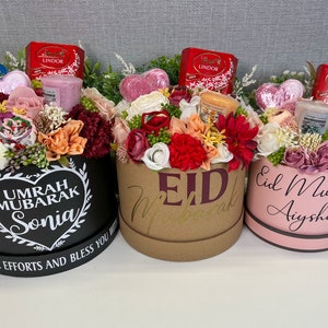 Pode incluir: Tr&ecirc;s caixas de presente cheias de flores, velas e chocolates. As caixas s&atilde;o decoradas com mensagens diferentes, incluindo "Umrah Mubarak Sonia", "Eid Mubarak" e "Eid Mubarak Aysha".