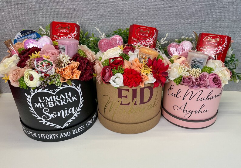 Pode incluir: Tr&ecirc;s caixas de presente cheias de flores, velas e chocolates. As caixas s&atilde;o decoradas com mensagens diferentes, incluindo "Umrah Mubarak Sonia", "Eid Mubarak" e "Eid Mubarak Aysha".