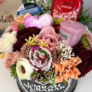 Pode incluir: Uma caixa de presente preta com um design de cora&ccedil;&atilde;o branco e o texto "UMRAH MUBARAK Sonia" cercada por uma variedade de flores artificiais, incluindo rosas cor-de-rosa, flores brancas e flores cor de p&ecirc;ssego. A caixa tamb&eacute;m cont&eacute;m uma bomba de banho em forma de cora&ccedil;&atilde;o rosa, uma pequena vela rosa e uma caixa de chocolates Lindor.