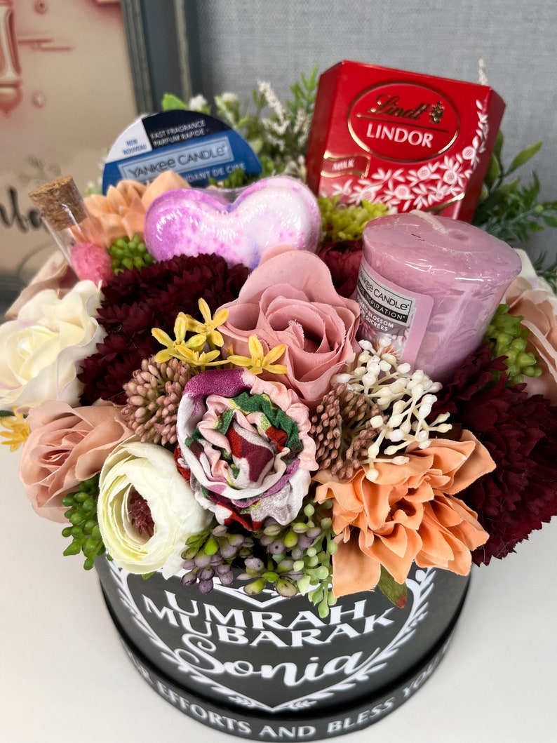 Pode incluir: Uma caixa de presente preta com um design de cora&ccedil;&atilde;o branco e o texto "UMRAH MUBARAK Sonia" cercada por uma variedade de flores artificiais, incluindo rosas cor-de-rosa, flores brancas e flores cor de p&ecirc;ssego. A caixa tamb&eacute;m cont&eacute;m uma bomba de banho em forma de cora&ccedil;&atilde;o rosa, uma pequena vela rosa e uma caixa de chocolates Lindor.