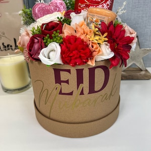 Pode incluir: Uma caixa de presente de cart&atilde;o castanho com as palavras "Eid Mubarak" em letras douradas e bord&ocirc;. A caixa est&aacute; cheia de uma variedade de flores artificiais, incluindo rosas vermelhas, brancas e cor-de-rosa, e uma pequena vela em forma de cora&ccedil;&atilde;o. H&aacute; tamb&eacute;m uma pequena caixa de chocolates Lindor e uma pequena vela.