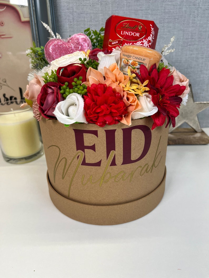 Pode incluir: Uma caixa de presente de cart&atilde;o castanho com as palavras "Eid Mubarak" em letras douradas e bord&ocirc;. A caixa est&aacute; cheia de uma variedade de flores artificiais, incluindo rosas vermelhas, brancas e cor-de-rosa, e uma pequena vela em forma de cora&ccedil;&atilde;o. H&aacute; tamb&eacute;m uma pequena caixa de chocolates Lindor e uma pequena vela.