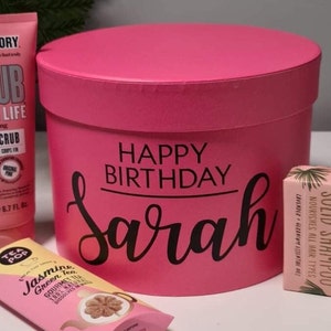 Pu&ograve; includere: Scatola regalo rosa con le parole "Happy Birthday Sarah" su di essa. La scatola contiene uno scrub corpo rosa, una barretta di shampoo solido e una bustina di t&egrave;.
