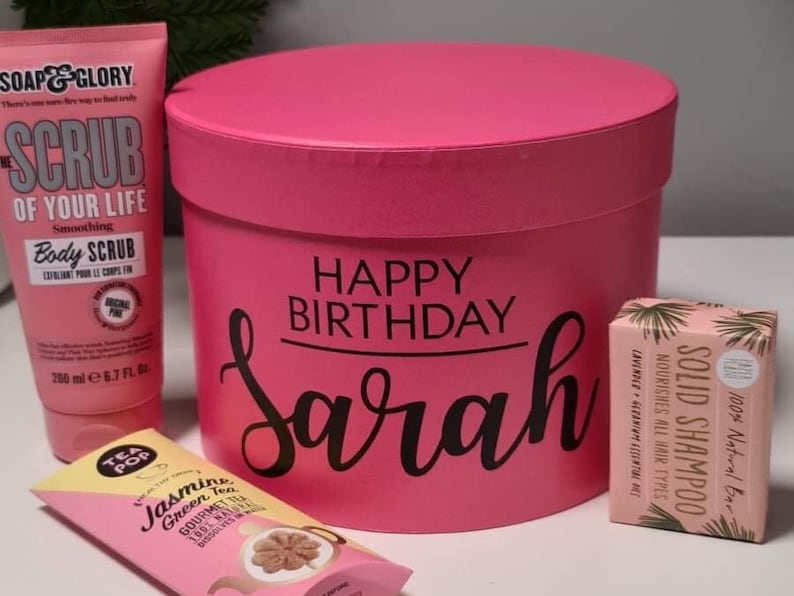 Pu&ograve; includere: Scatola regalo rosa con le parole "Happy Birthday Sarah" su di essa. La scatola contiene uno scrub corpo rosa, una barretta di shampoo solido e una bustina di t&egrave;.