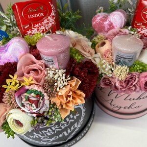Pode incluir: Duas caixas de presente redondas cheias de flores, velas e chocolate. As caixas s&atilde;o decoradas com as palavras "Umrah Mubarak Sonia" e "Love Aysha".