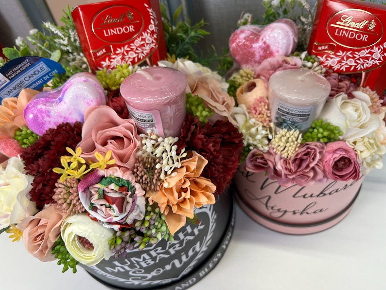 Pode incluir: Duas caixas de presente redondas cheias de flores, velas e chocolate. As caixas s&atilde;o decoradas com as palavras "Umrah Mubarak Sonia" e "Love Aysha".