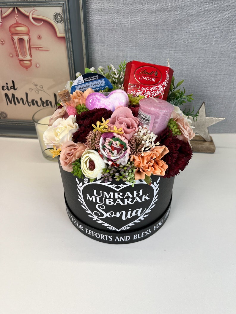 Pode incluir: Uma caixa de presente preta com um design de cora&ccedil;&atilde;o branco e o texto "Umrah Mubarak Sonia" cercada por um arranjo floral. A caixa cont&eacute;m uma vela rosa, uma bomba de banho em forma de cora&ccedil;&atilde;o, uma pequena caixa de chocolates e um difusor de fragr&acirc;ncia.
