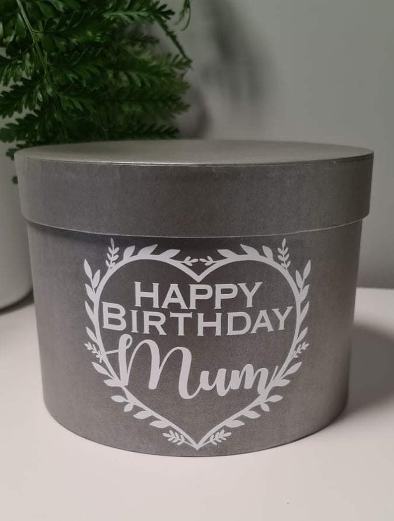 Pu&ograve; includere: Una scatola regalo rotonda grigia con un design a forma di cuore bianco e il testo "Happy Birthday Mum" in una ghirlanda di foglie.