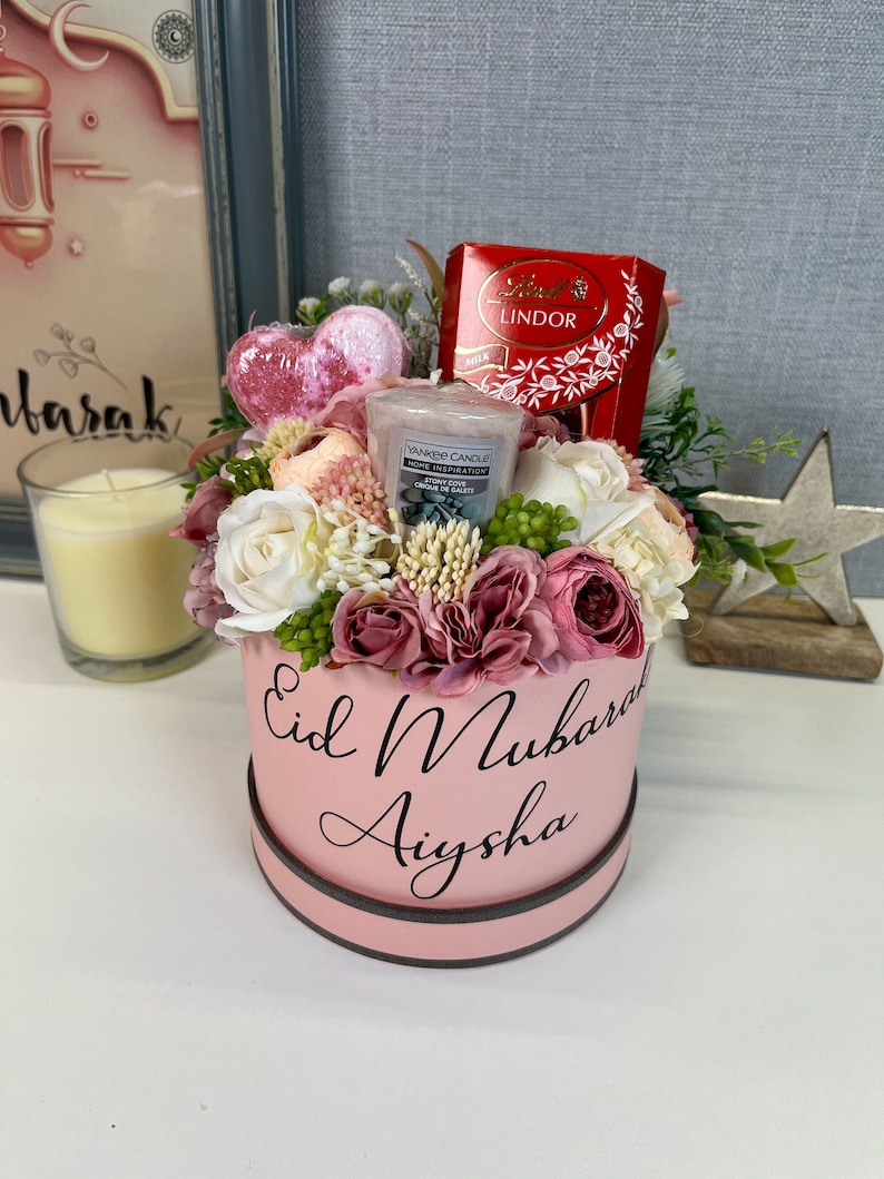 Pode incluir: Uma caixa de presente rosa com as palavras "Eid Mubarak Aysha" nela. A caixa est&aacute; cheia de flores rosa e brancas, uma vela em forma de cora&ccedil;&atilde;o, uma vela pequena e uma caixa de chocolates Lindor.
