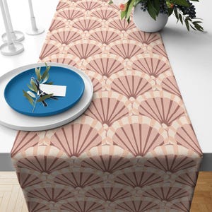 May include: A table runner featuring a repeating fan-shaped shell pattern in shades of dusty rose, light pink, and cream. The fabric is laid flat on a surface, partially covered by a white plate topped with a blue plate and a small sprig of greenery.