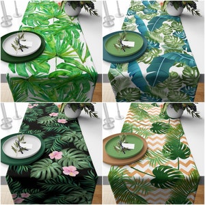 Può includere: Quattro diversi runner da tavolo con motivi di foglie tropicali. Ogni runner presenta un diverso motivo di foglie verdi e bianche. I runner sono mostrati su un tavolo con una mise en place su ciascuno.