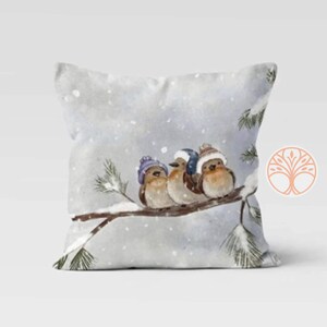 Three birds wearing purple and blue knit hats are perched on a snowy pine branch. The birds are brown with white bellies and are looking to the right. The background is a watercolor painting of a snowy winter scene.