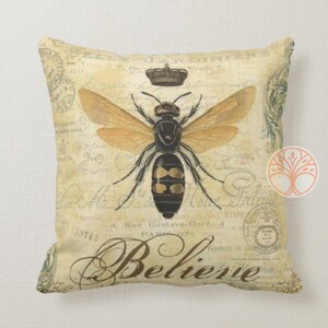 May include: A decorative throw pillow with a vintage design featuring a bee with wings spread, a crown, and the word "Believe" in a script font. The background is a cream color with a distressed look and includes French text.