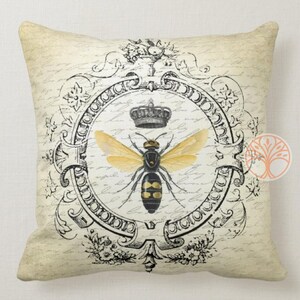 May include: A decorative throw pillow with a vintage design featuring a black and yellow bee with gold wings inside a black and white ornate frame. The bee is wearing a crown. The background is a cream color with black script writing.