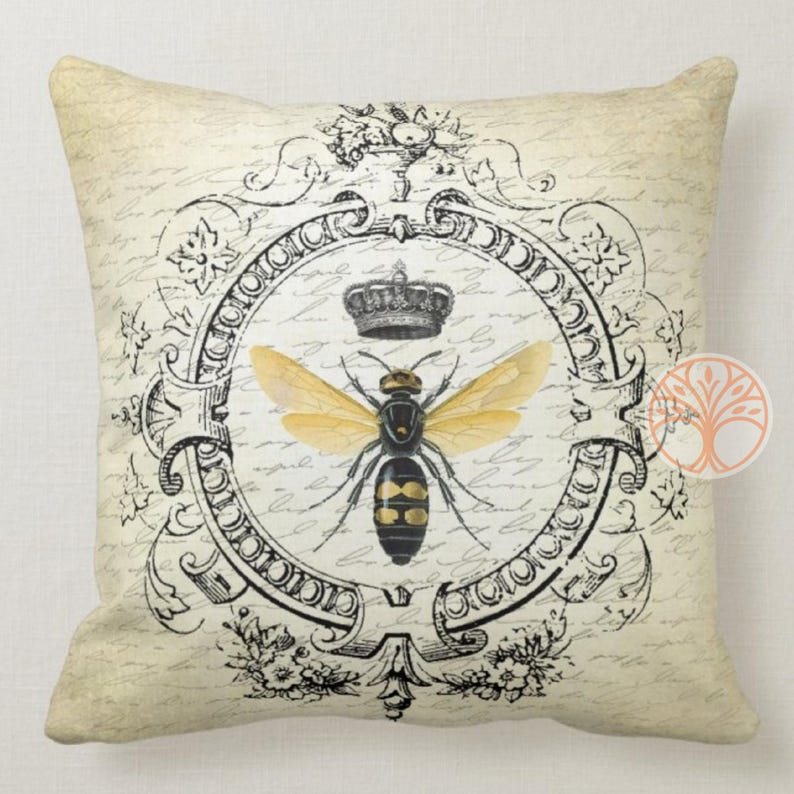 May include: A decorative throw pillow with a vintage design featuring a black and yellow bee with gold wings inside a black and white ornate frame. The bee is wearing a crown. The background is a cream color with black script writing.