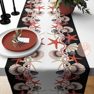 May include: A white table runner with black borders features a repeating pattern of seashells, starfish, and coral. The runner is laid out on a table with a red plate and olive branches.