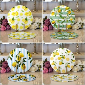 May include: Four round ceramic plates with different lemon designs. The plates have a white background with yellow lemons and green leaves. The plates are on a table with flowers and a vase.