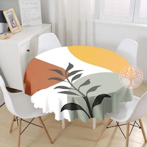 May include: A round white tablecloth with a geometric design featuring abstract shapes in shades of orange, yellow, and gray. A black leafy branch design is centered on the tablecloth.