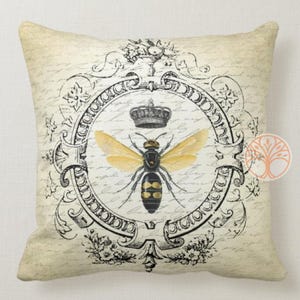 May include: A decorative throw pillow with a vintage design featuring a black and yellow bee with gold wings inside a black and white ornate frame. The bee is wearing a crown. The background is a cream color with black script writing.