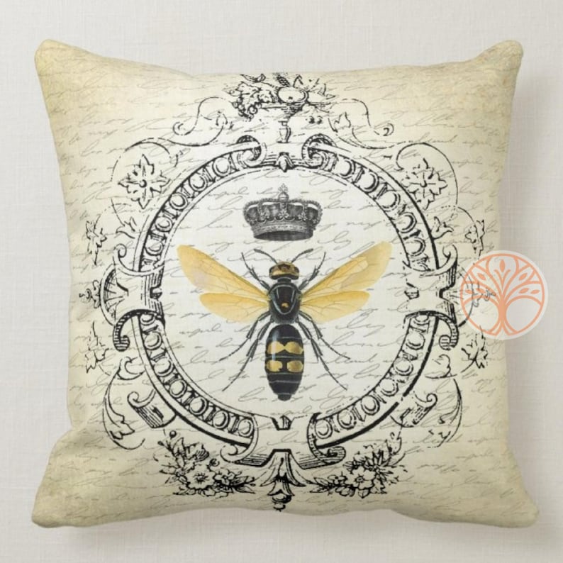 May include: A decorative throw pillow with a vintage design featuring a black and yellow bee with gold wings inside a black and white ornate frame. The bee is wearing a crown. The background is a cream color with black script writing.