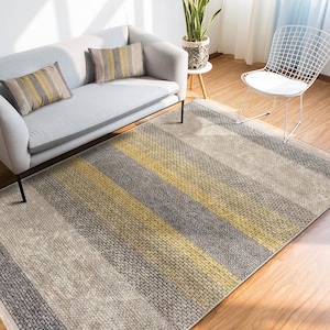 Könnte beinhalten: Ein rechteckiger Teppich mit Streifenmuster in Grau-, Beige- und Gelbtönen. Der Teppich liegt auf einem Holzboden, im Hintergrund ein graues Sofa und Kissen. Das Design des Teppichs zeigt abwechselnde Farbstreifen.