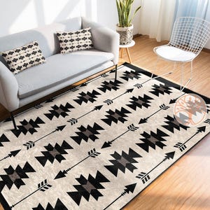 May include: A black and white geometric rug with a pattern of arrows and squares. The rug is on a wooden floor and there is a white couch and a chair in the background.