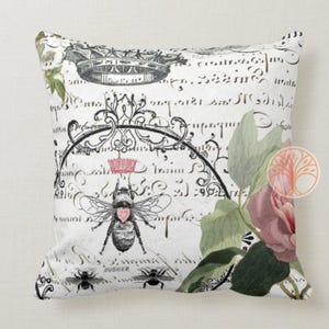May include: A white pillow with a black and white floral design featuring a bee with a pink heart. The background is a white and black script.