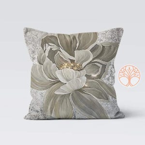 A decorative throw pillow with a large, white flower with a gold center on a gray background. The flower is stylized and has a soft, watercolor-like appearance.