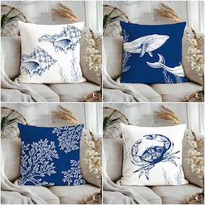 Może przedstawiać: Zestaw czterech kwadratowych poduszek dekoracyjnych z motywami marynistycznymi. Poduszki przedstawiają białe i granatowe ilustracje muszli, wielorybów, koralowców i kraba. Poduszki leżą na neutralnej kanapie.