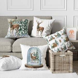 May include: Five decorative throw pillows with winter forest and deer designs. The pillows are on a light brown couch with a wicker basket in the foreground. The pillows are white with watercolor illustrations of deer, snow-covered trees, and a cabin. The text "AKASIA" is visible on a framed picture behind the pillows.