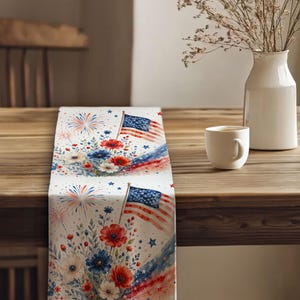 May include: A white table runner featuring a watercolour print of American flags, red and blue flowers, and firework bursts. The design includes scattered blue stars and red and blue swashes, creating a patriotic pattern across the fabric surface.