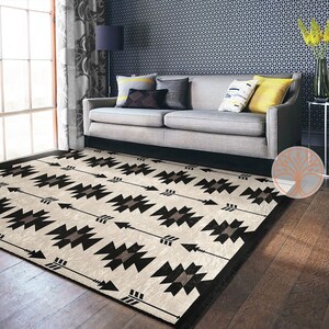 May include: A black and white geometric patterned rug with a cream background. The rug features a repeating pattern of arrows and geometric shapes. The rug is laid out on a hardwood floor in front of a gray couch.