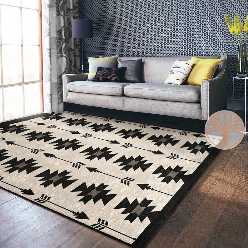 May include: A black and white geometric patterned rug with a cream background. The rug features a repeating pattern of arrows and geometric shapes. The rug is laid out on a hardwood floor in front of a gray couch.