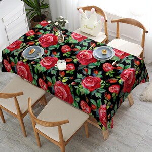Könnte beinhalten: Ein rechteckiges Tischtuch mit einem auffälligen Blumenmuster aus großen roten Rosen und grünen Blättern auf schwarzem Hintergrund. Der Tisch ist mit Tellern, Besteck und einer weißen Tasse gedeckt. Zwei Holzstühle sind sichtbar.