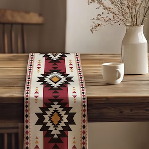 May include: A decorative table runner with a Southwestern geometric pattern. The runner features a central burgundy stripe with black, yellow, and white geometric shapes. The edges have a repeating pattern of triangles and diamonds. A white mug and vase are on the wooden table.