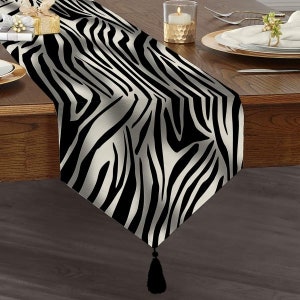May include: A black and white zebra print table runner with a black tassel. The runner is on a wooden table with a candle and a plate.