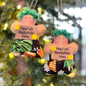 May include: Two handmade felt ornaments shaped like people wearing green leis and floral print skirts. Each ornament holds a sign that reads "Hau'oli Makahiki Hou" which is Hawaiian for "Happy New Year".