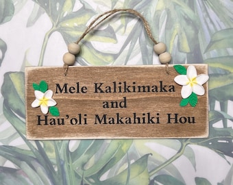 Op de afbeelding: Houten bord met de tekst "Mele Kalikimaka and Hau'oli Makahiki Hou" in zwarte letters. Het bord is versierd met witte en gele bloemaccenten en een jute touw om op te hangen. De achtergrond heeft een tropisch bladerpatroon.