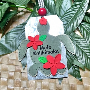 Op de afbeelding: Een decoratief schildpadvormig ornament met de tekst "Mele Kalikimaka". Het ornament is grijs met rode bloemaccenten en groene bladeren. Het wordt opgehangen met een zwart-wit geruit lint en een rode kraal.