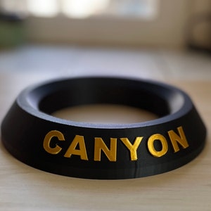 May include: A black ring-shaped object with the word "CANYON" written in gold letters.