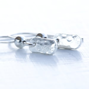 May include: A pair of silver hoop earrings with a textured rectangular charm hanging from each hoop.