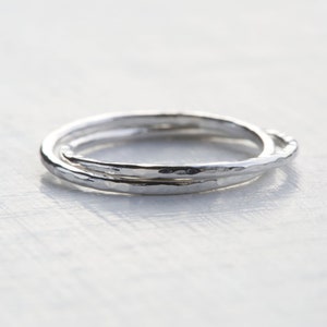 Puede incluir: Dos anillos de plata, uno ligeramente más grande que el otro, están entrelazados y tienen una textura martillada. Los anillos están sobre una superficie blanca.