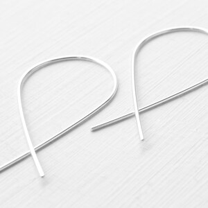Op de afbeelding: Een paar minimalistische zilveren oorbellen. Elke oorbel heeft een gebogen, langwerpig ontwerp, dat lijkt op een slanke, open lus. De oorbellen zijn gemaakt van een glanzend, metalen materiaal en staan tegen een witte achtergrond.