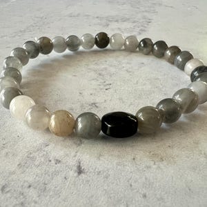 Könnte beinhalten: Ein graues, weißes und braunes Perlenarmband mit einer schwarzen ovalen Perle in der Mitte.
